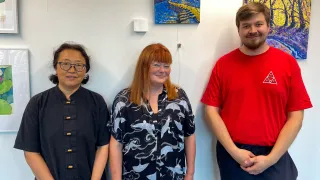 Three art club members standing in front of their art and smiling at the camera.