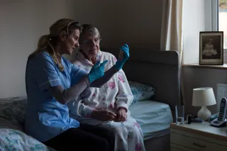 A nurse and senior person are sitting side by side on a bed, looking at a test sample that the nurse is holding